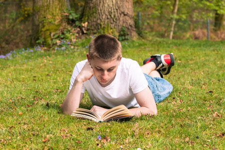 Teenage boy reading a book in the sun in a public park. Wearing a white T-shirt and blue denimの写真素材