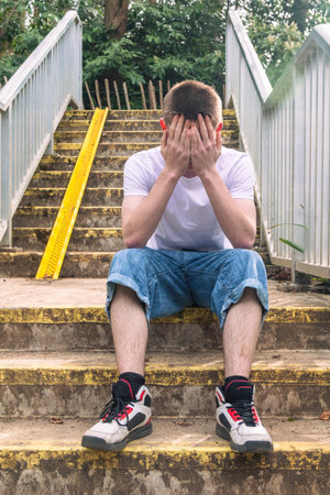 Depressed Teenage boy with his head in his hands, in white t-shirt and blue denim sitting on stairs outside in a public parkの写真素材