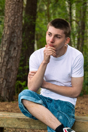 Happy Teenage boy sititng on a bench in a public park, wearing a white t-shirt and blue denimの写真素材