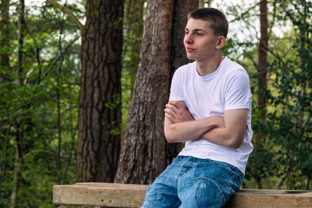 Teenage boy sititng on a bench in a public park, wearing a white t-shirt and blue denimの写真素材