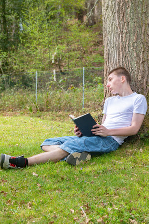 Teenage boy resting against a tree while reading a bookの写真素材