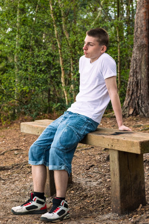 Teenage boy sititng on a bench in a public park, wearing a white t-shirt and blue denimの写真素材