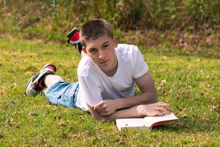 Teenage boy reading a book in the sun in a public park. Wearing a white T-shirt and blue denimの写真素材