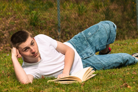 Teenage boy reading a book in the sun in a public park. Wearing a white T-shirt and blue denimの写真素材