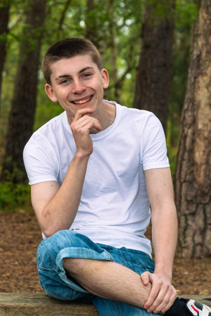 Happy Teenage boy sititng on a bench in a public park, wearing a white t-shirt and blue denimの写真素材