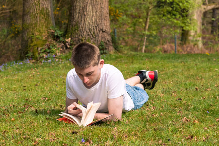 Teenage boy reading a book in the sun in a public park. Wearing a white T-shirt and blue denimの写真素材