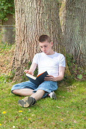 Teenage boy resting against a tree while reading a bookの写真素材