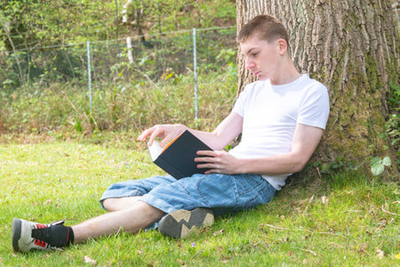Teenage boy resting against a tree while reading a bookの写真素材