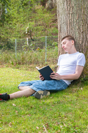 Teenage boy resting against a tree while reading a bookの写真素材