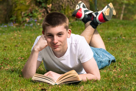 Teenage boy reading a book in the sun in a public park. Wearing a white T-shirt and blue denimの写真素材