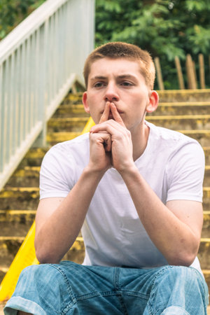 Thoughtful Teenage with his fingers on his lips, boy in white t-shirt and blue denim sitting on stairs outside in a public parkの写真素材