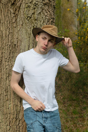Teenage boy wearing a bush hat, white t-shirt and blue denim. On hand is on the hat, pulling it downの写真素材