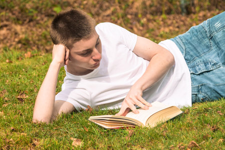 Teenage boy reading a book in the sun in a public park. Wearing a white T-shirt and blue denimの写真素材