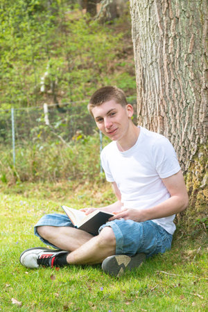 Teenage boy sitting against a tree while reading a bookの写真素材
