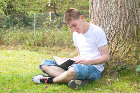 Teenage boy sitting against a tree while reading a bookの写真素材