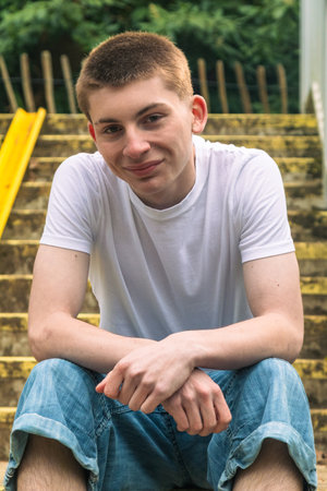 Happy Teenage boy with his chin on his hand, in white t-shirt and blue denim sitting on stairs outside in a public parkの写真素材