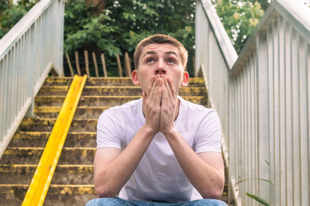 Teenage boy looking up, in white t-shirt and blue denim sitting on stairs outside in a public parkの写真素材
