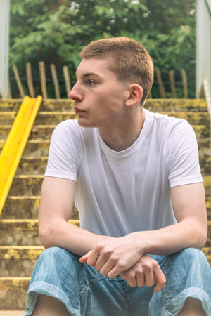 Serious Looking Teenage boy in white t-shirt and blue denim sitting on stairs outside in a public parkの写真素材