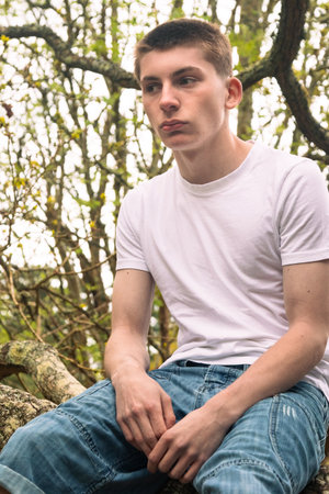 A teenage boy sitting in a tree in a public park, wearing a white t-shirt and blue denimの写真素材