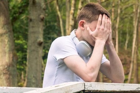 Teenage boy wearing white t-shirt and blue denim leaning on wooden railings outside. with headphones round his neckの写真素材