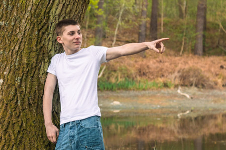 Teenage boy wearing a white t-shirt and blue denim, leaning on a tree, near a canal in a public parkの写真素材