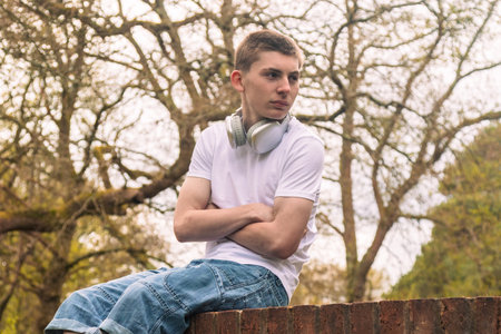 Teenage boy wearing a white t-shirt and blue denim sitting on a brick wall with headphones round his neckの写真素材