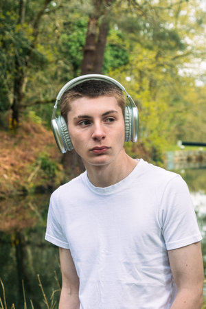 Teenage boy wearing a white t-shirt and blue denim, outside in a public park near a canal listening to music via headphonesの写真素材