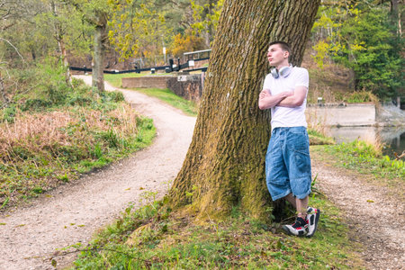 Teenage boy wearing a white t-shirt and blue denim, outside in a public park, leaning against a treeの写真素材