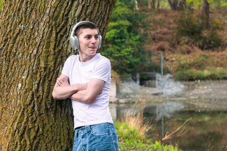 Teenage boy wearing a white t-shirt and blue denim, outside in a public park, leaning against a tree,  near a canal listening to music via headphonesの写真素材