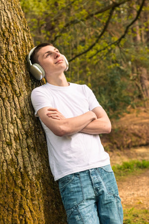 Teenage boy wearing a white t-shirt and blue denim, outside in a public park near a canal, leaning against a tree listening to music via headphonesの写真素材