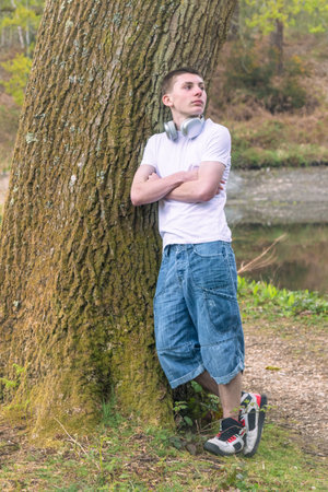 Teenage boy wearing a white t-shirt and blue denim, outside in a public park, leaning against a tree, near a canal with headphones round his neckの写真素材