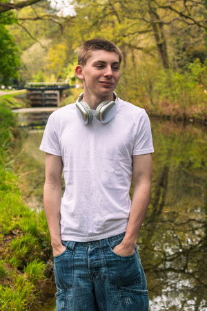 Teenage boy wearing a white t-shirt and blue denim, outside in a public park near a canal with headphones round his neckの写真素材