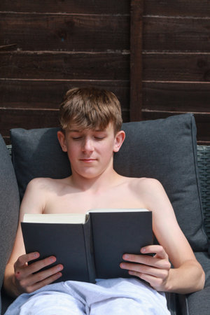 A shirtless teenage boy reading a book on an outdoors sofa on a summer's dayの写真素材