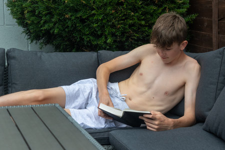 A shirtless teenage boy reading a book on an outdoors sofa on a summer's dayの写真素材