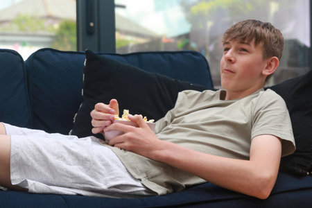 A Teenage Boy Laying On A Sofa With A Bowl of Popcorn While Watching TVの写真素材