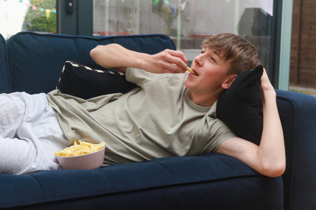 A Teenage Boy Laying On A Sofa Eating A Bowl Of Potato Snacks While Watching TVの写真素材