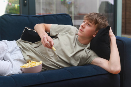 A Teenage Boy Laying On A Sofa Eating A Bowl Of Potato Snacks While Watching TVの写真素材