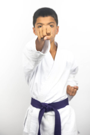 A portrait of a preteen British boy of Iranian heritage wearing a traditional white karate gi doing a head punch with selective focus