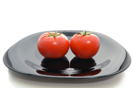 Two fresh tomatoes with green stems on a black plate isolated on white backgroundの写真素材