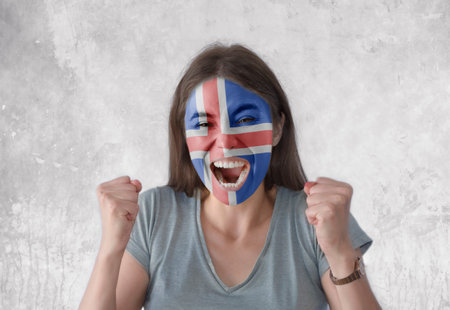 Young woman with painted flag of Iceland and open mouth looking energetic with fists upの写真素材
