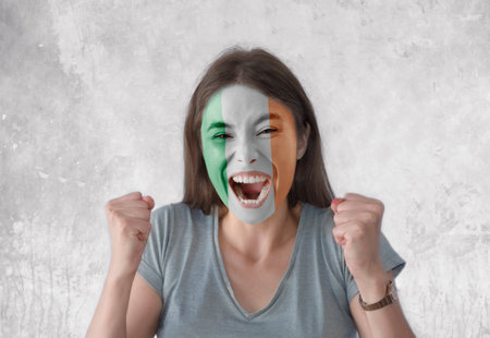 Young woman with painted flag of Ireland and open mouth looking energetic with fists upの写真素材