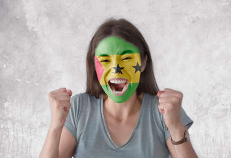 Young woman with painted flag of Sao Tome and Principe and open mouth looking energetic with fists upの写真素材
