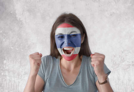 Young woman with painted flag of Thailand and open mouth looking energetic with fists upの写真素材