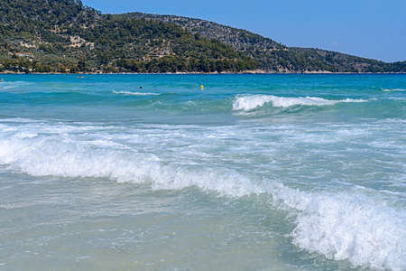 Big wave of ocean on sandy beach with blue clean sky and green hillの写真素材
