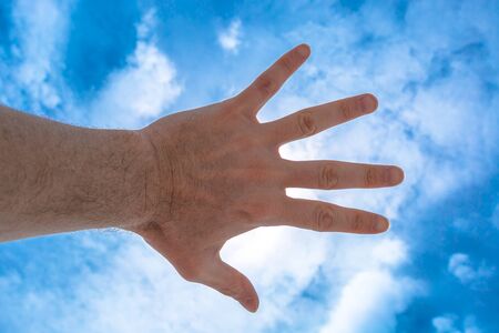 Hand of a man reaching out against a cloudy blue sky, covering the sun. Concept of business and communication of self-confidence. Concept of focus on yourself and clear vision of the problems.の写真素材