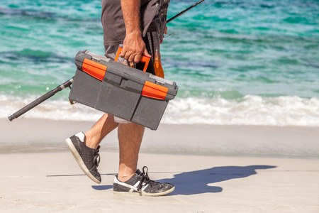 Man walking on the beach holding in one hand a tackle box and a fishing rod in the other.の写真素材
