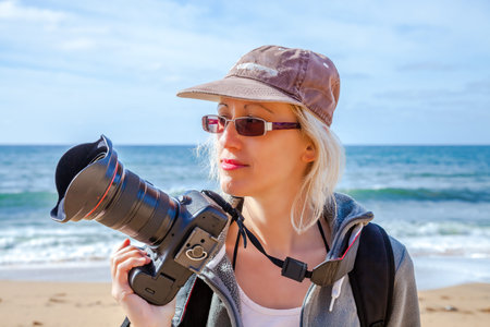 Woman professional photographer taking pictures on a beach, freehand. Sea landscape background.
