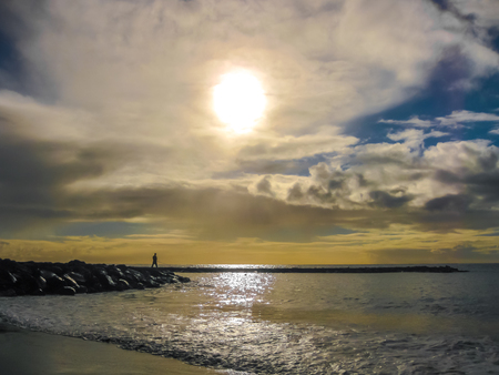 Fisherman on the rocks with a fishing rod in his hand. Beach Las Americas, Tenerife, Canary Islands. Cloudy sky at sunset with sun rays that pass through the clouds.の写真素材