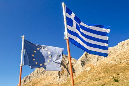 Flags of Greece and Europe waving on the blue sky with mountains in the background.