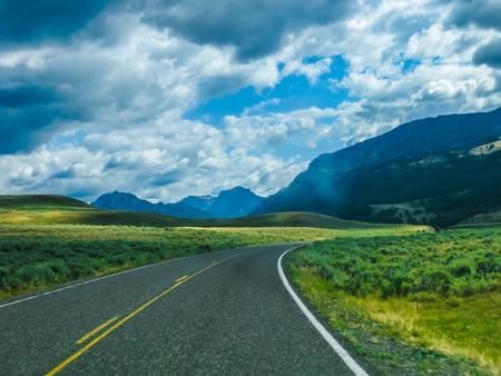 Paved road in the Yellowstone National Park, Wyoming, United States, between prairies, mountains and cloudy sky.の写真素材